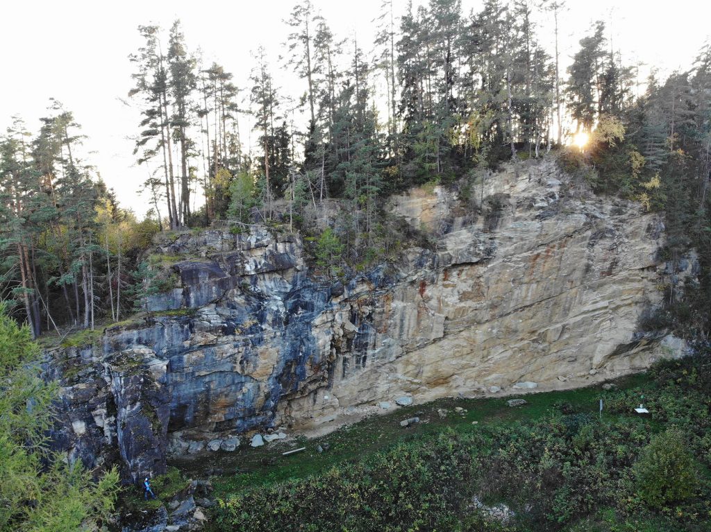 Das Bild zeigt den Klettergarten Seebichl-Kraig von der Luft aus, der gelbliche Fels ragt über einer Wiese hervor, dahinter Bäume und die untergehende Sonne.