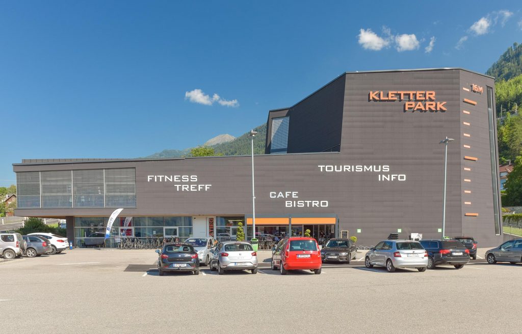 Das Bild zeigt eine der größten Kletterhallen Kärnten, die Halle in Mühldorf. Ein graues Gebäude, parkende Autos vor blauem Himmel.
