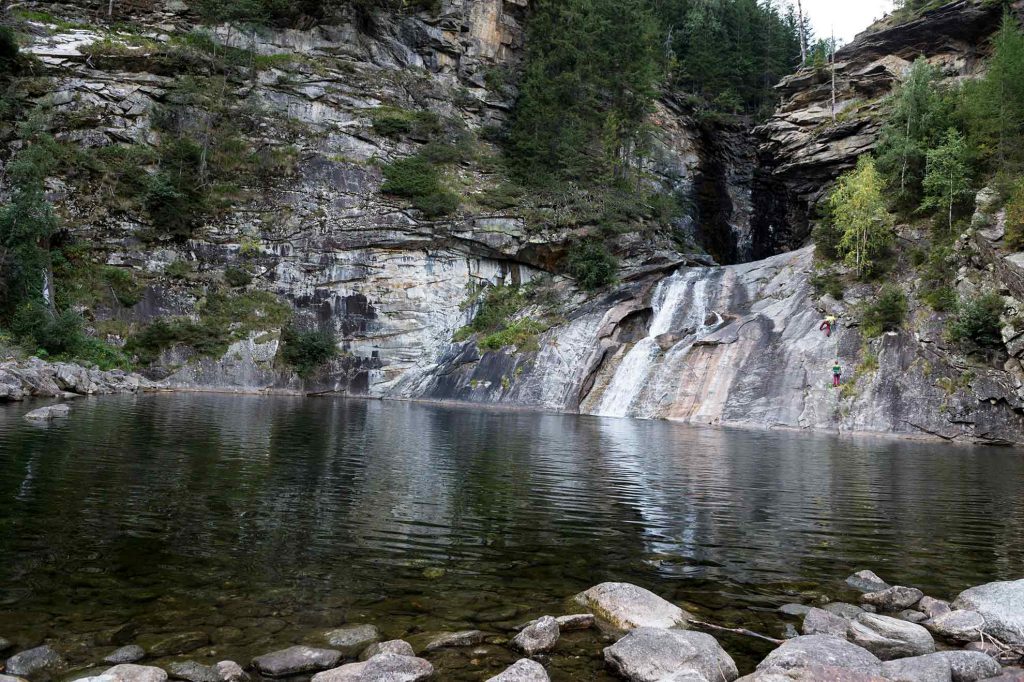 Das Bild zeigt den Blauen Tumpf im Maltatal. Hinter einer großen, kristallklaren Wasserfläche stürzt ein kleiner Wasserfall in das Becken. Rechts und links braune Felsen.