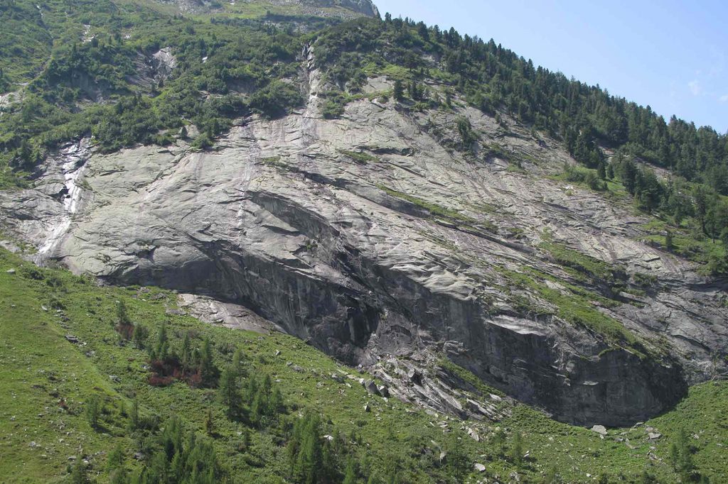 Das Bild zeigt einen breiten Granit Panzer mit einem zarten Wasserfall in der Mitte. Darüber Wald, darunter grüne Wiese.