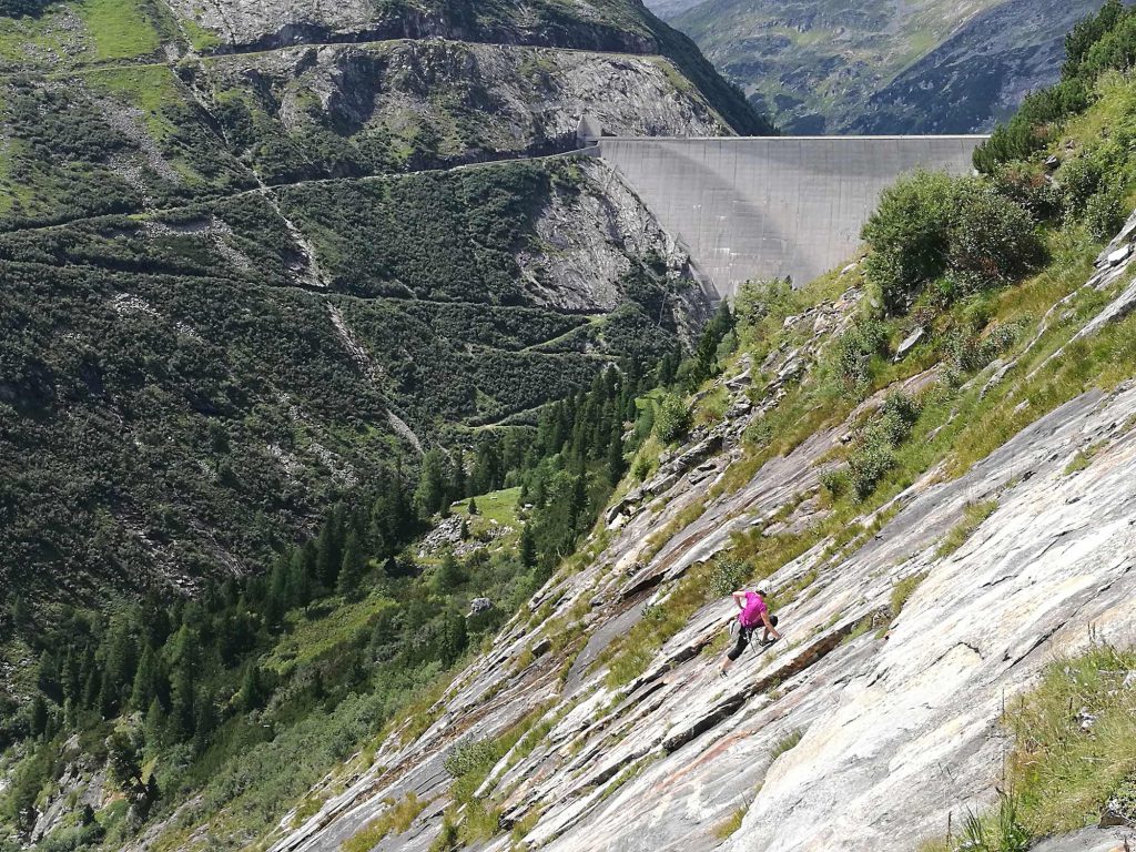 Das Bild zeigt eine Klettererin auf einer großen Platte vor der Kölnbreinsperre Staumauer und dem gegenüberliegenden Berghang.