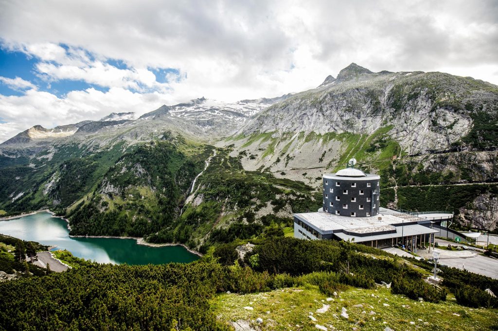 Das Bild zeigt die Berglandschaft rund um den Klettersteig Kölnbreinsperre. Eine beeindruckende Berglandschaft, im Vorderteil des Bildes ein Hotel mit rundem Gebäude, dahinter felsige Landschaft, einen See und schneebedeckte Berge.