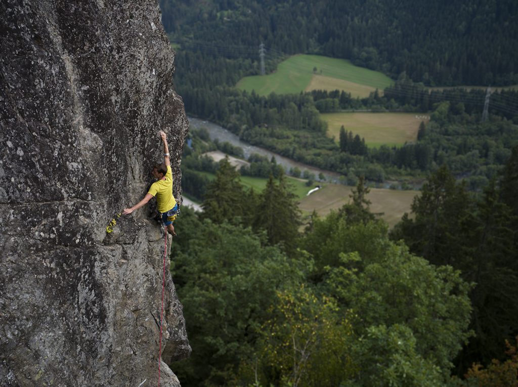 Das Bild zeigt einen der Klettergärten im Mölltal, den Danielsberg. Ein Kletterer mit gelbem Shirt klettert an einer Kante, im Hintergrund Bäume sowie der grüne Talboden und Felder.