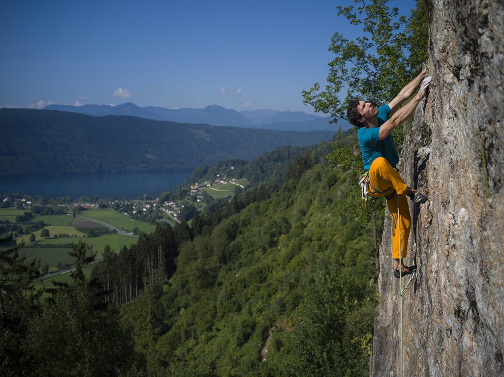 Das Bild zeit einen Kletterer mit oranger Hose und türkisem Shit in der Breitwand hoch über dem Millstätter See.