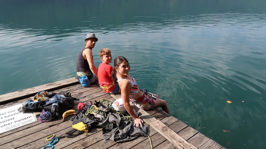 Das Bild zeigt eine Gruppe von Jungen Leuten im Klettergarten Jungfernsprung am Millstätter See. Sie sitzen auf einem Floß mit den Füßen im Wasser und lächeln in die Camera.