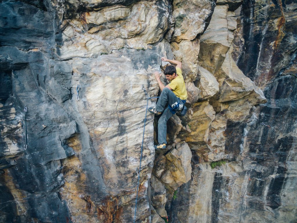 Das Bild zeigt einen Kletterer im Klettergarten Kraig - Seebichl. Er setzt dynamisch zu einer Bewegung an einer Felskante an. Der Fels in seiner unmittelbaren ist gelb, darum herum grauer Fels. Er trägt eine graue Hose und ein gelbes T-Shirt.