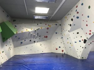 Das Bild zeigt eine der Kletterhallen Kärnten, nämlich die Naturfreunde Boulderhalle Klagenfurt. Auf grauen Kletterwänden sind bunte Griffe, darunter eine blaue Niedersprungmatte.