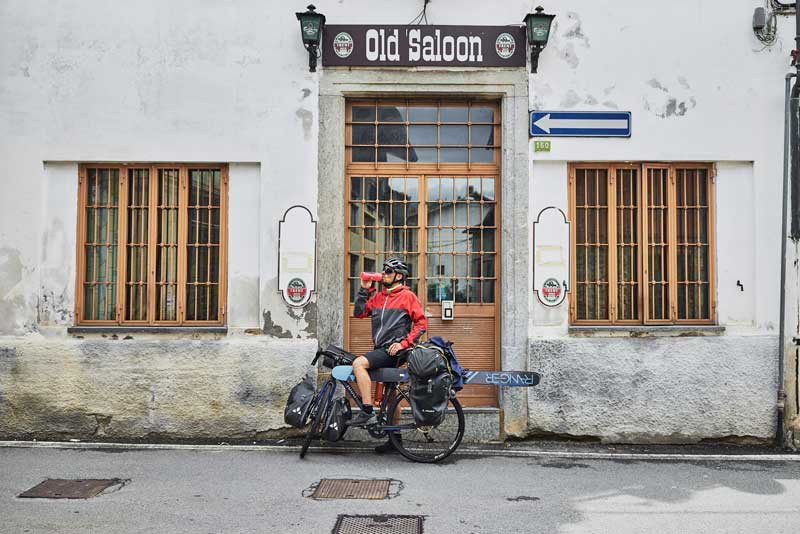 Das Bild zeigt einen Radfahrer in Regenkleidung vor einem Old Saloon. Am Rad hat er Freeride Skier befestigt.