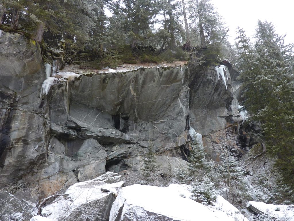 Das Bild zeigt den Drytooling Sektor Wingmaster, einen wichtigen Teil des Eisklettern Maltatal. Ein grauer, winterlicher Felsen mit Eiszapfen, darüber Wald und greller Himmel.