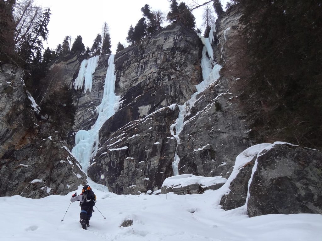 Das Bild zeigt eine der Locations beim Eisklettern Maltatal, die Eisfälle Odin und Hidden Treasure. In einer grauen Felswand ziehen zwei Eisfälle separat empor. Auf der Felskante und seitlich Bäume, oben greller Himmel.