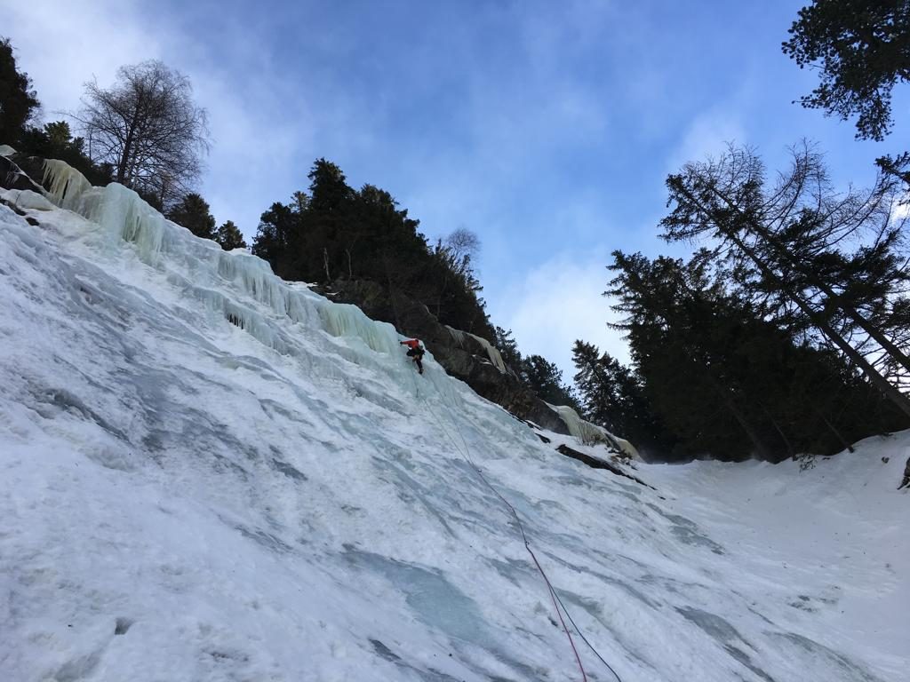 Das Bild zeigt das Eisklettern Maltatal, einen Eiskletterer im Maaralmfall. Im Bild Vordergrund die breite Eisfront des Wasserfalles, weit oben ein Eiskletterer. An der Felskante dunkle Bäume, darüber blauer Himmel.