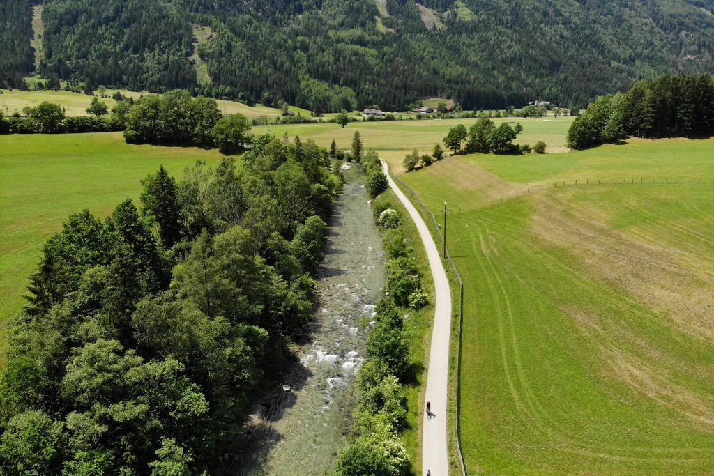 Das Bild zeigt den Radweg durch das Maltatal beim Ort Feistritz von einer Drohne aus. Zwischen grünen Wiesen fließt der Malta Fluss, parallel dazu verläuft der graue Radweg. Am unteren Bildrand sieht man klein zwei Radfahrer.