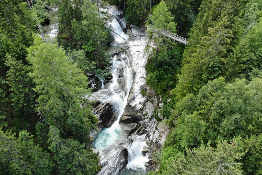 Das Bild zeigt den dritten der Gössfälle Maltatal mit der Aussichtsplattform und der Hängebrücke aus der Luft. Durch einen Wald zieht sich eine Schlucht mit Wasserfällen. Daneben grauer Fels und rundherum grüner Wald.