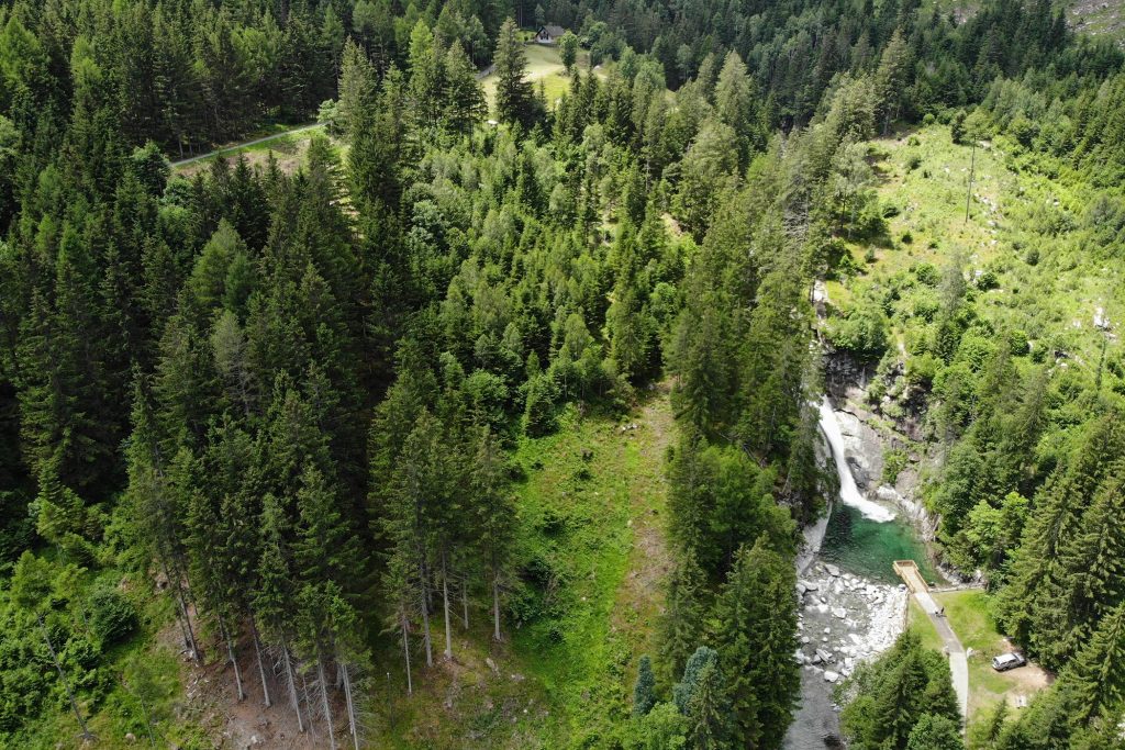 Das Bild zeigt die Gössfälle Maltatal beim 1. Gössfall. Mitten im Wald sieht man von einer Drohne aus den Wasserfall mit seinem grün-blauen Wasserbecken und die Aussichtsplattform. Darum herum alles grüner Wald.