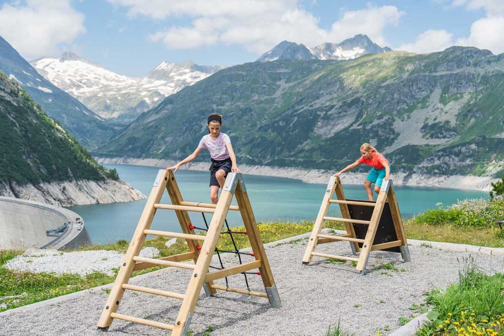 Zwei Mädchen am Abenteuerweg Kölnbrein auf sprossenböcken, im Hintergrund der STausee.