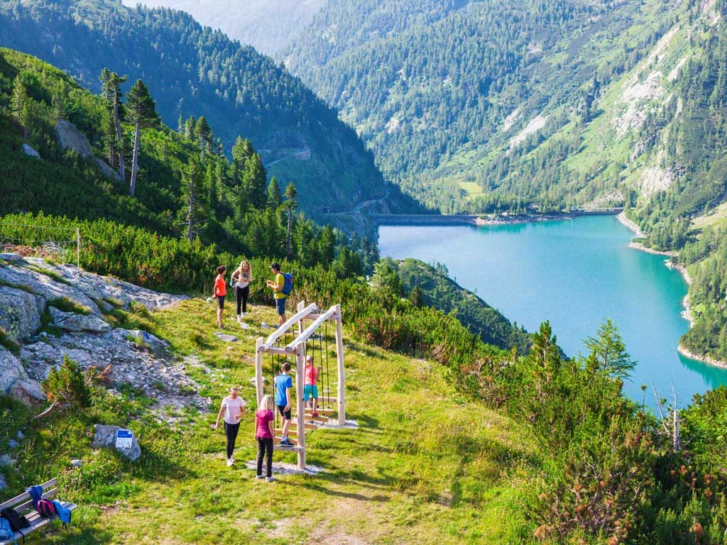 Gruppe mit Erwachsenen und Kindern am Erlebniswanderweg Kölnbrein, im Hintergrund ein Stausee und Berge.