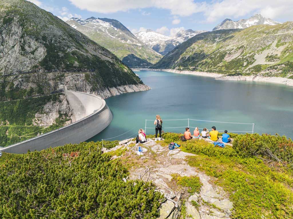 Eine Familie rastet auf einer Granitkuppe am Abenteuerweg Kölnbrein. Im Hintergrund der Stausee und alpine Landschaft.